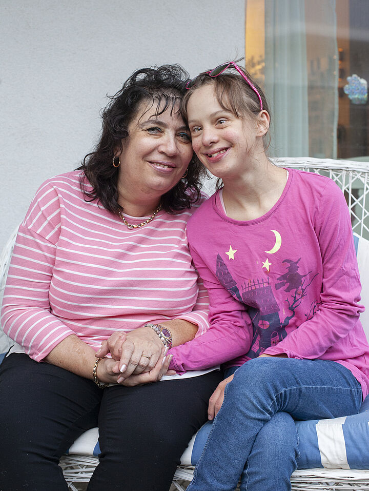Eine Mutter im rot-weiß gestreiften Oberteil und schwarzer Hose hält die Hand ihrer Tochter, die blaue Jeans und ein pinkes Shirt trägt.