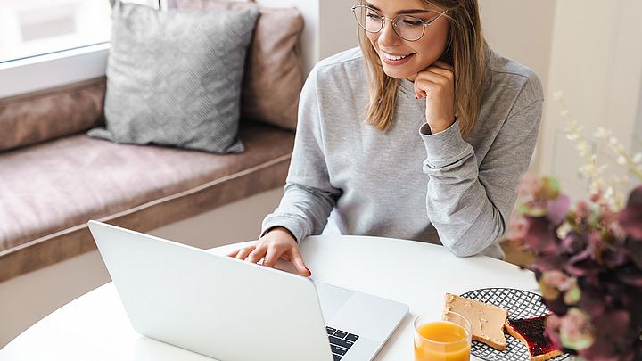 Eine junge Frau arbeitet zu Hause am Laptop während sie frühstückt.