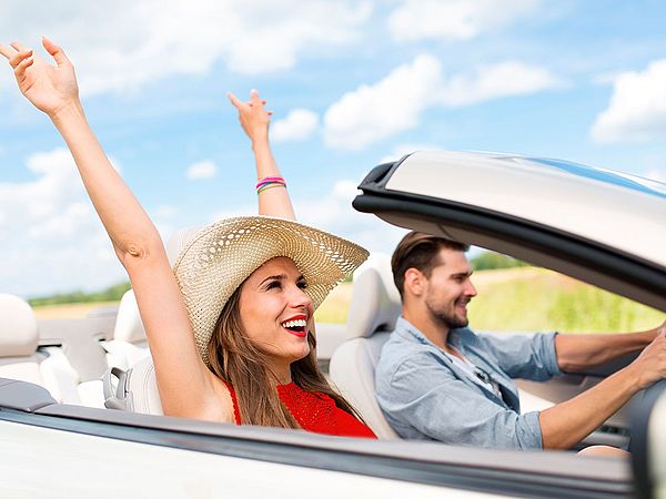 Ein junges glückliches Paar fährt gemeinsam in einem Cabrio. Die Frau streckt vor Freude ihre Arme in die Luft.