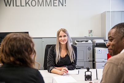 Blonde Dame sitzt an einem Schreibtisch. Ihr gegenüber sitzen ein Mann und eine Frau. Sie unterhalten sich.