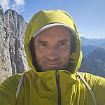 Selfie von einem Mann in gelb-grüner Jacke vor einer Bergkulisse mit Felswand im Hintergrund. Er hat die Kapuze aufgesetzt.