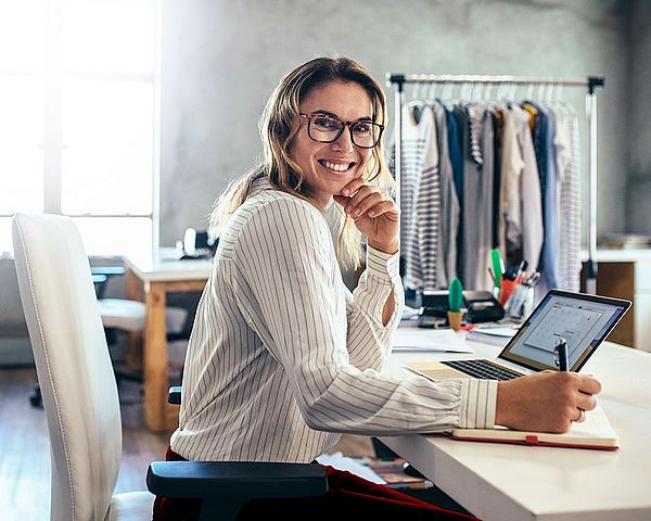 Eine Frau sitzt am Laptop im Büro eines Modeunternehmens. Sie lächelt dabei und schreibt Notizen auf einen Notizblock 