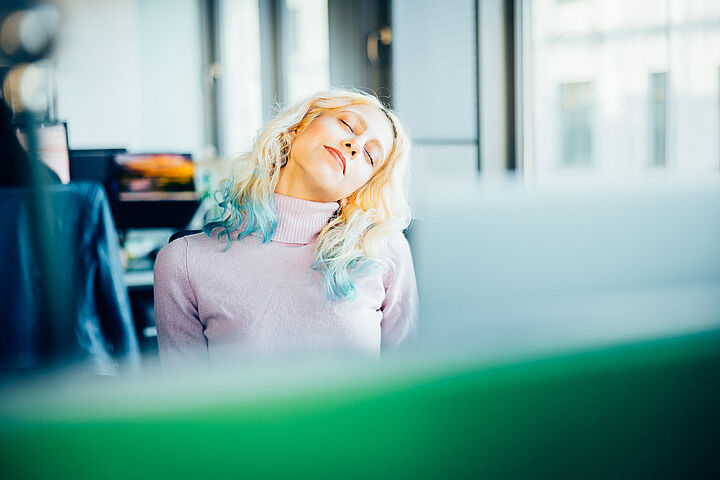 Eine junge Frau mit blond-türkisen Haaren schließt die Augen und legt ihren Kopf zur Seite. Sie trägt einen hellrosa Rollkragenpullover.