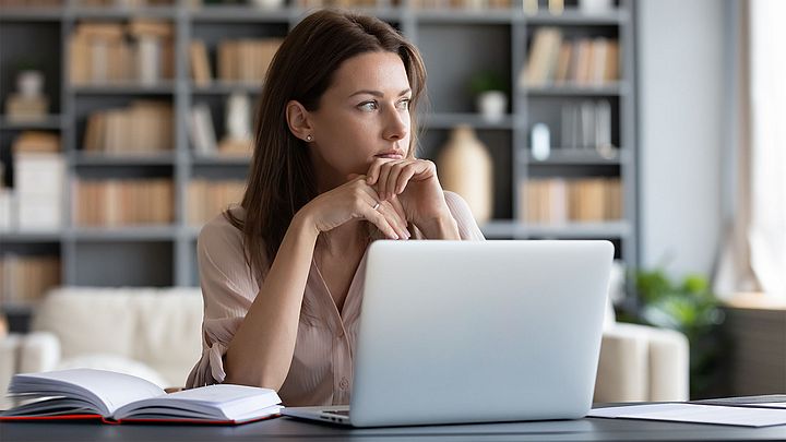 Junge Frau sitzt beim Schreibtisch mit ihrem Laptop und schaut nachdenklich aus.