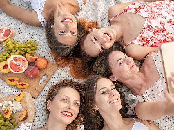 Eine Gruppe junger Frauen macht ein Picknick im Freien. Dabei nehmen sie ein Selfie auf während sie Kopf an Kopf am Boden liegen 