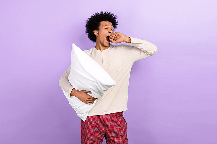 Ein junger Mann gähnt: er hat ein Kopfpolster unter dem Arm und eine rotkarierte Pyjamahose an.