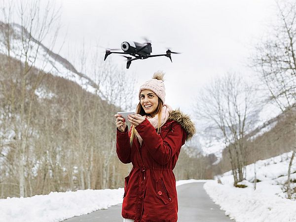 Eine junge Frau steuert lächelnd eine Drohne in einer Winterlandschaft.