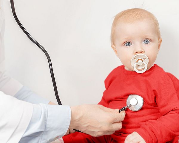 Ein kleines Mädchen mit einem Schnuller im Mund schaut in Kamera, während die Ärztin es mit einem Stethoskop untersucht.