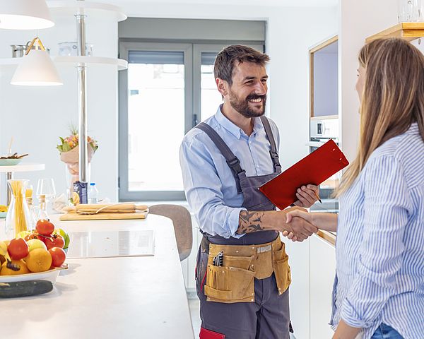 Ein Handwerker mit Latzhose schüttelt lächelnd die Hand einer Frau. Dabei befinden sie sich in einer Küche.