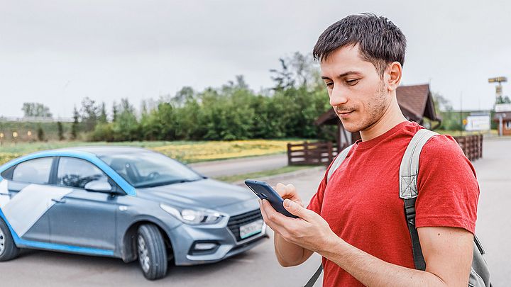 Ein junger Mann steht auf einem Parkplatz neben einem Auto und tippt etwas in sein Smartphone.