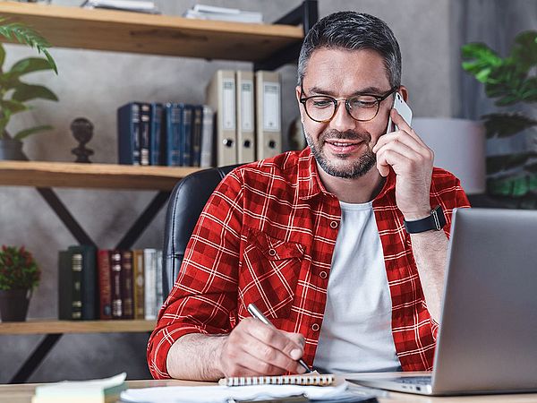 Ein Mann sitzt am Laptop im Büro und schreibt etwas auf einem Notizblock während er telefoniert.