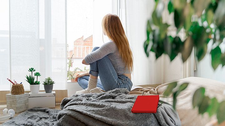 Eine junge Frau sitzt auf ihrem Bett und schaut aus dem Fenster.