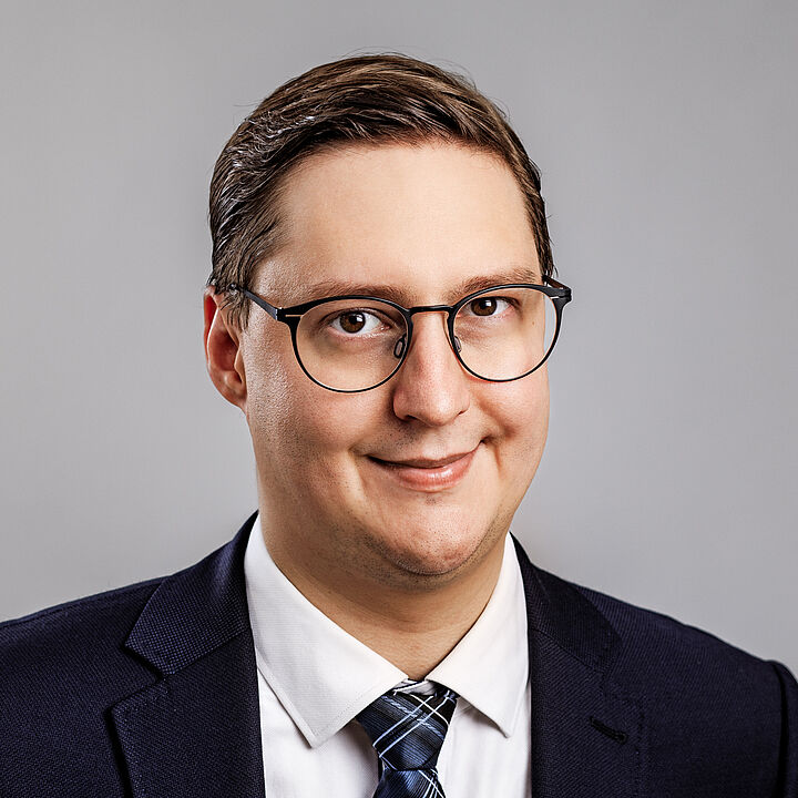 Porträtbild eines jungen Mannes mit Brille, schwarzem Anzug und Krawatte. Er hat kurzes Haar und lächelt.