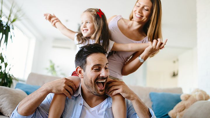 Eine glückliche Familie ist zu Hause im Wohnzimmer. Die Tochter sitzt auf den Schultern ihres Vaters.