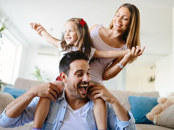 Eine glückliche Familie ist zu Hause im Wohnzimmer. Die Tochter sitzt auf den Schultern ihres Vaters.