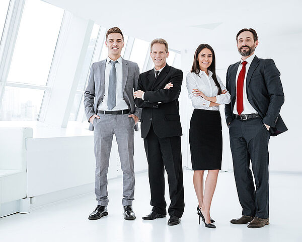 Eine Gruppe von Geschäftsleuten steht in einem Büro zusammen und schauen dabei lächelnd in die Kamera