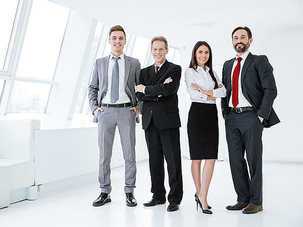 Eine Gruppe von Geschäftsleuten steht in einem Büro zusammen und schauen dabei lächelnd in die Kamera