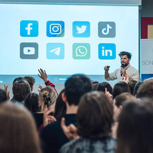 Ein Mann steht vor einer großen Leinwand und spricht mit einem Mikrofon in der Hand zu einem Publikum. Auf der Leinwand werden acht verschiedene Social-Media-Icons (wie Facebook, Instagram, TikTok) angezeigt.