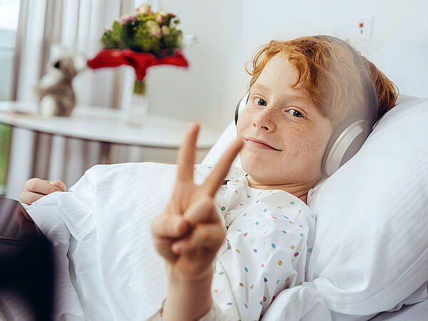Junge liegt in Spitalsbett mit Kopfhörern. Er lächelt und zeigt mit den Fingern das Victory Symbol.