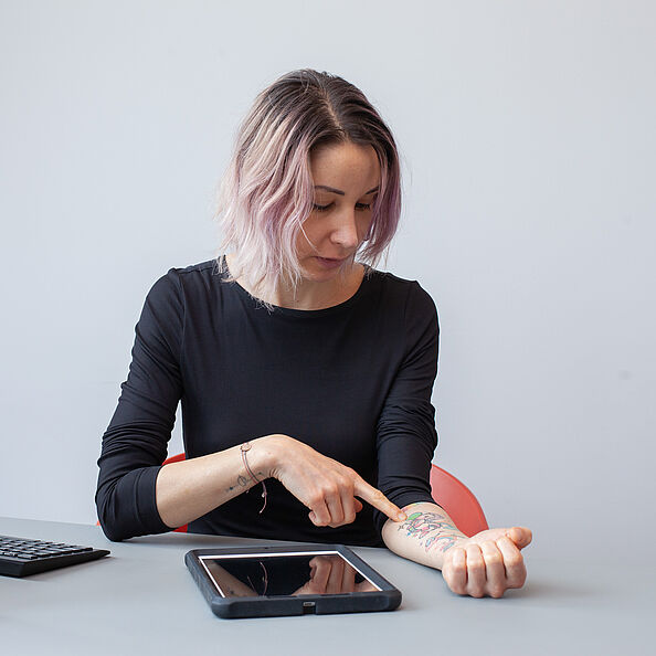 Eine junge Frau mit lila Haaren sitzt an einem Schreibtisch, legt ihren Unterarm auf den Tisch und zeigt auf eines ihrer Tattoos am Unterarm.
