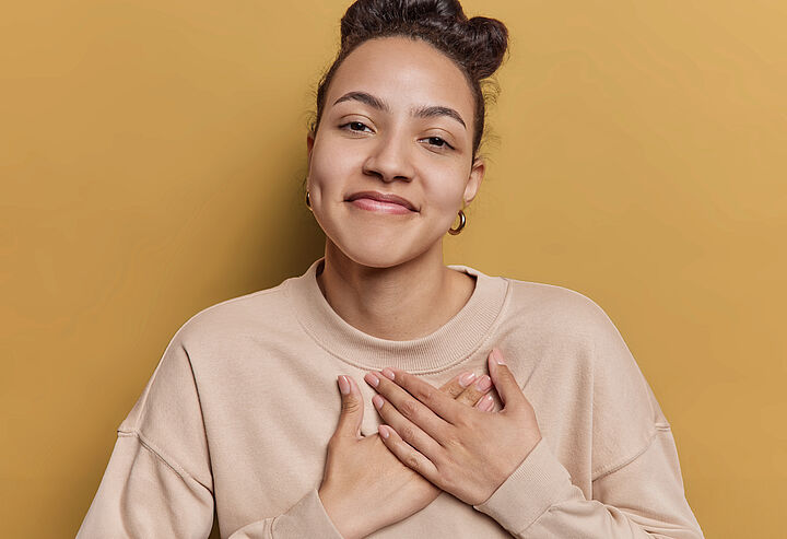 Eine junge Frau im beigen Pullover mit dunklen, hochgesteckten Haaren lächelt und hält beide Hände auf ihr Herz.