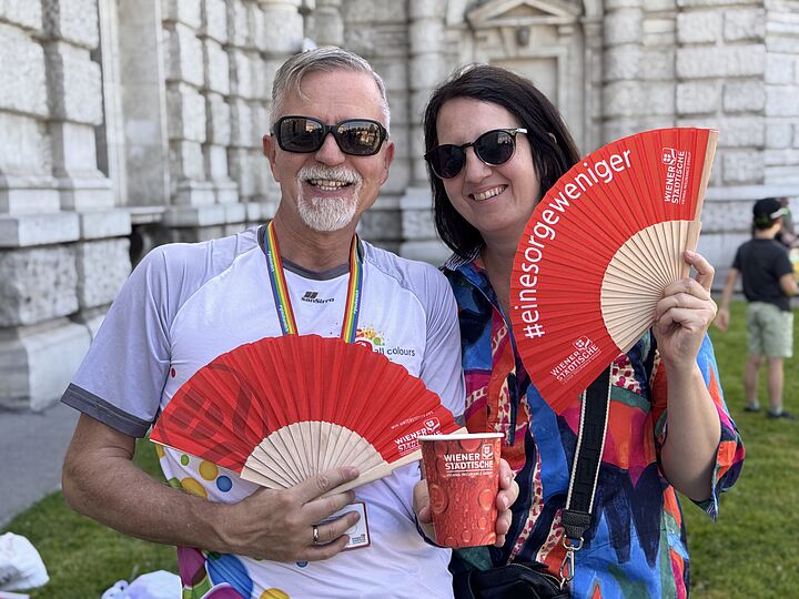 Zwei Personen mit Sonnenbrille, die einen Fächer in der Hand halten und in die Kamera lachen.