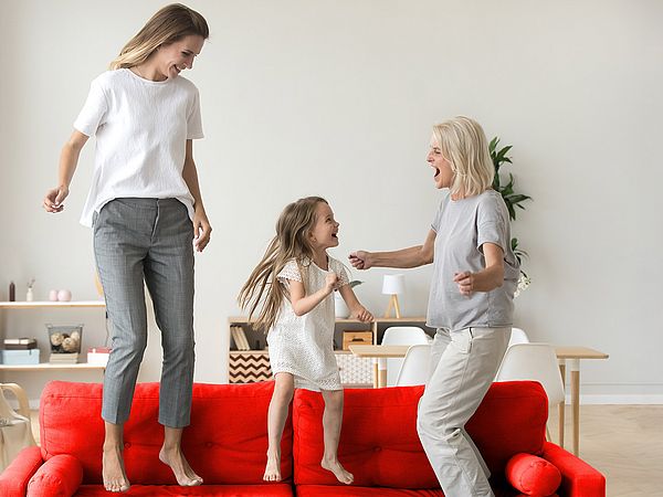 Ein glückliches junges Mädchen springt mit ihrer Mutter und ihrer Großmutter auf dem Sofa. 