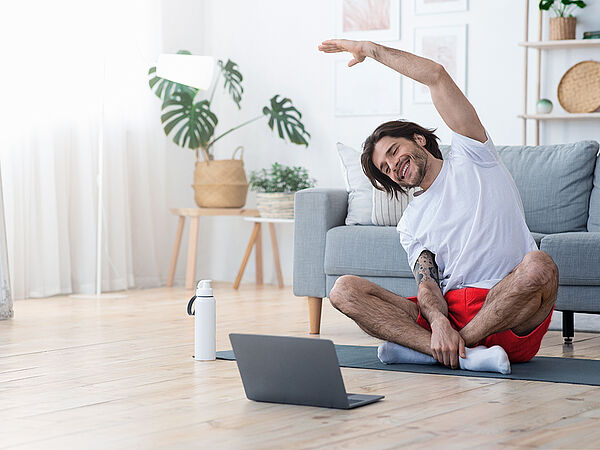 Ein junger Mann macht Gymnastikübungen zu Hause auf seiner Yoga-Matte vor seinem Laptop