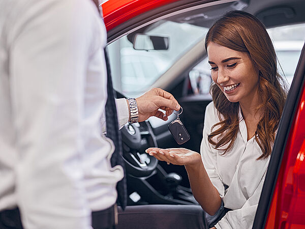 Eine glückliche junge Frau sitzt in ihrem neuen Auto und nimmt den Autoschlüssel vom Verkäufer entgegen.