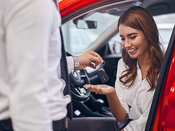 Eine glückliche junge Frau sitzt in ihrem neuen Auto und nimmt den Autoschlüssel vom Verkäufer entgegen.