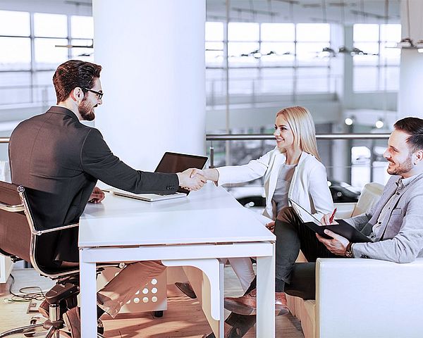 Ein Geschäftsmann sitzt mit Kunden im Büro. Dabei schüttelt er der Kundin die Hand, während der andere Kunde lächelnd dabei zusieht 