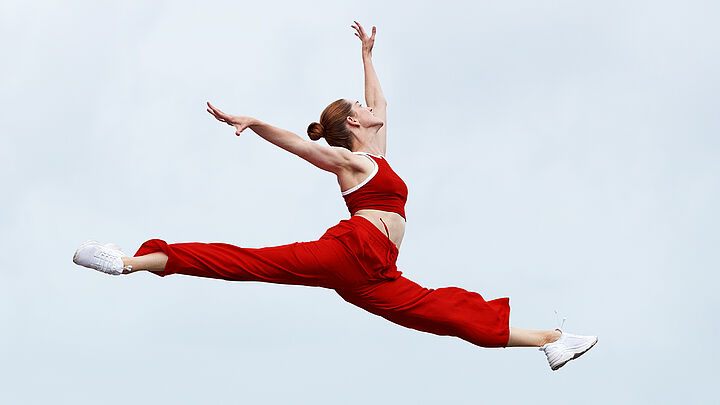 Junge blonde Frau in rotem Sportoutfit macht einen hohen Spagatsprung.