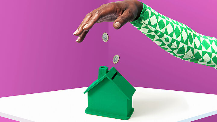 Studioaufnahme einer Hand, die zwei Euromünzen in eine grüne Spardose wirft, die wie ein kleines Haus aussieht