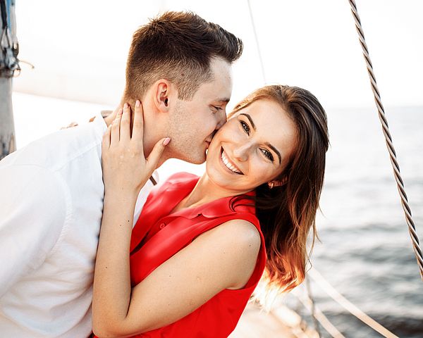 Ein glückliches junges Paar entspannt auf einem Boot am Meer. Der Mann gibt der Frau einen Kuss auf die Wange.