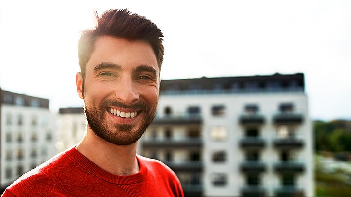 Ein junger Mann steht im Freien in einer Stadt und lächelt dabei in die Kamera 