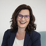 Lachende Frau mit dunklem Sakko und weißem Shirt mit Brille.