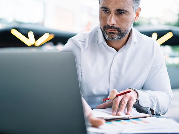 Ein Geschäftsmann sitzt am Schreibtisch vor seinem Laptop und macht sich konzentriert Notizen 