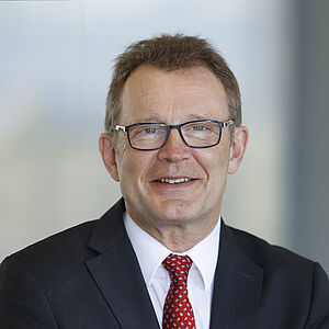 Porträtbild von einem Herrn mit dunkler Brille, schwarzem Sakko, weißem Hemd und roter Krawatte.