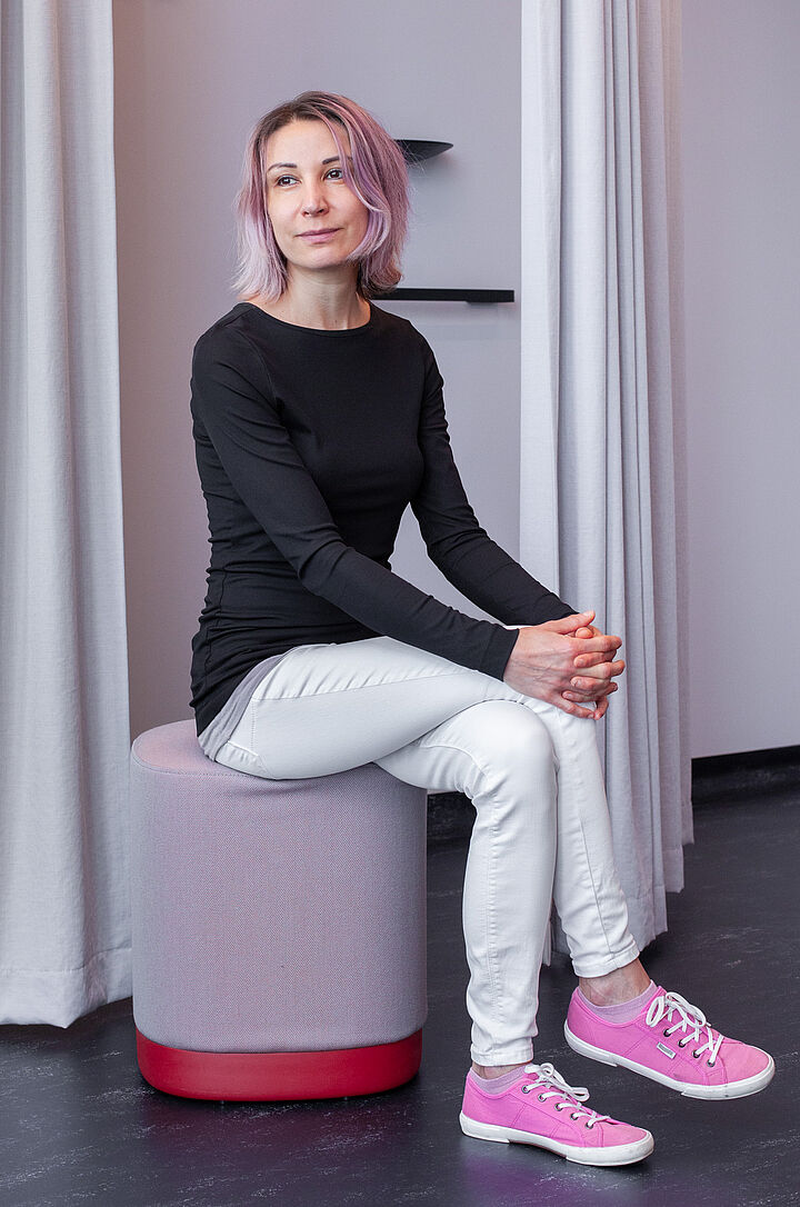 Eine junge Frau mit lila Haaren sitzt auf einem gepolsterten Hocker. Sie trägt eine helle Hose, ein schwarzes, langärmliges Oberteil und pinke Sneakers.