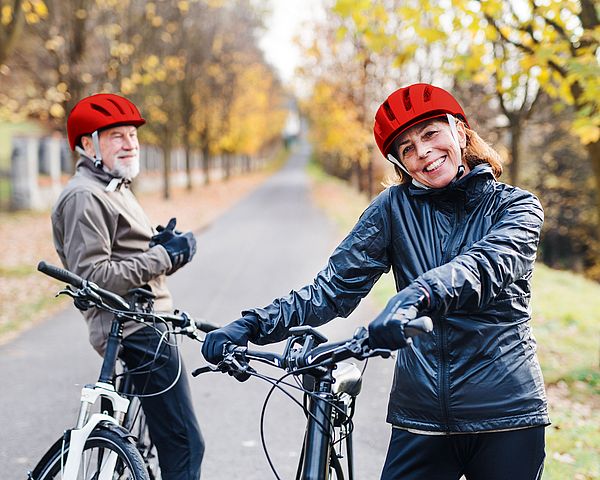 Ein glückliches Seniorenpaar in Sportbekleidung steht mit Fahrrädern auf einer Straße in der Natur.