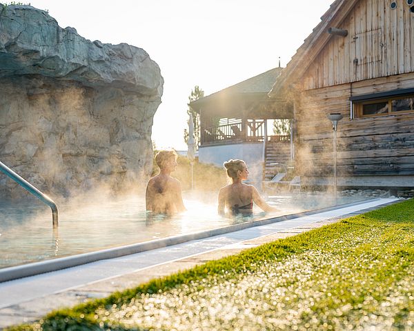 Mann und Frau schwimmen im warmen Thermalwasser umgeben von Wiesen und Holzhütten.