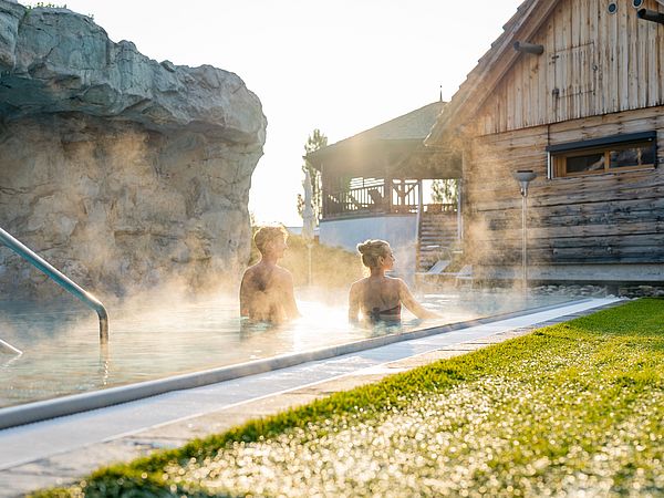 Mann und Frau schwimmen im warmen Thermalwasser umgeben von Wiesen und Holzhütten.