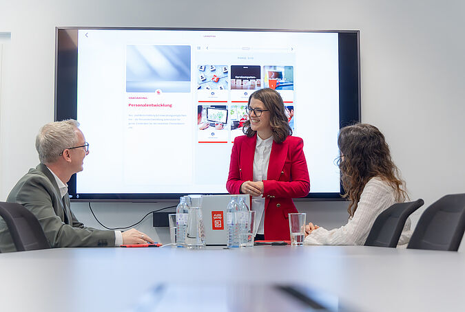 Eine Frau in rotem Hosenanzug steht vor einem Smartboard in einem Besprechungsraum und spricht mit einem Mann und einer Frau.