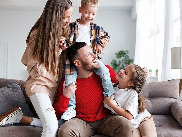 Eine junge glückliche Familie sitzt zu Hause auf der Couch. Der Sohn sitzt auf den Schultern seines Vaters und die beiden Töchter sitzen neben ihm