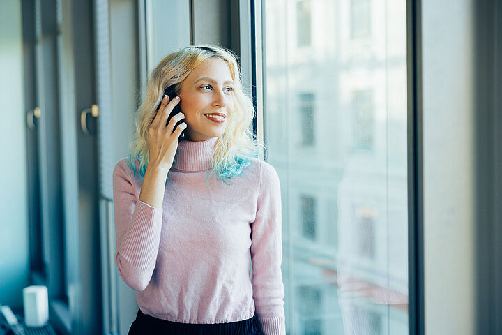 Eine junge Frau mit blond-türkis gefärbten Haaren im rosa Rollkragenpullover steht lächelnd am Fenster und telefoniert mit ihrem Smartphone.