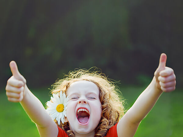 Ein Mädchen mit einer Blume im lockigen Haar befindet sich auf einer Wiese und streckt voller Freude beide Daumen nach oben.