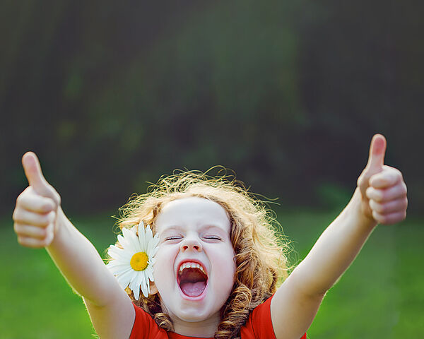 Ein Mädchen mit einer Blume im lockigen Haar befindet sich auf einer Wiese und streckt voller Freude beide Daumen nach oben.