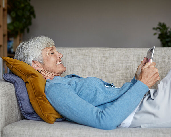 Ältere Frau mit kurzen grauen Haaren liegt entspannt auf einem Sofa und lächelt, während sie ein Smartphone in den Händen hält.