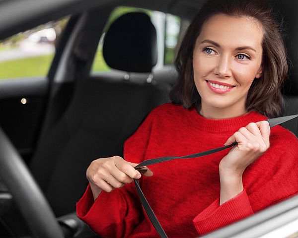Eine lächelnde junge Frau sitzt am Fahrersitz und schnallt sich an.