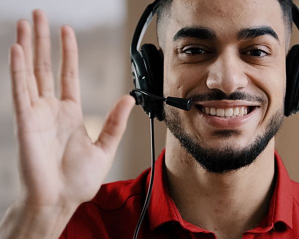 Ein Mann mit Headset lächelt in die Kamera und winkt dabei